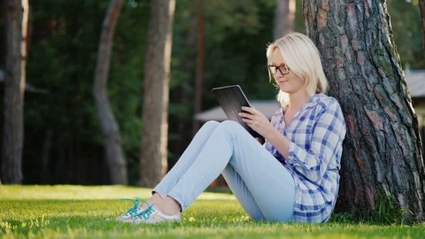 A young woman is using a tablet. Sits on the grass under a tree in the backyard Stock Footage 80049246