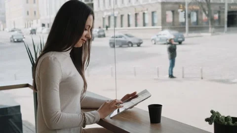 Young woman using a tablet sitting in cafe near the big window and watching city Stock Footage 205551902