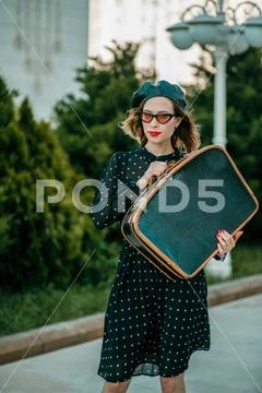 Young woman in vintage black polka dot dress with retro suitcase in hand posi Stock Photos