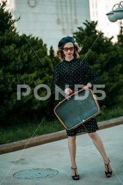 Young woman in vintage black polka dot dress with retro suitcase in hand posi Stock Photos