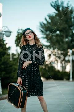 Young woman in vintage black polka dot dress with retro suitcase in hand posi Stock Photos