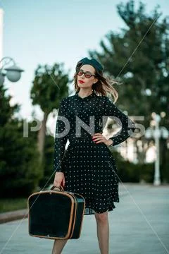 Young woman in vintage black polka dot dress with retro suitcase in hand posi Stock Photos