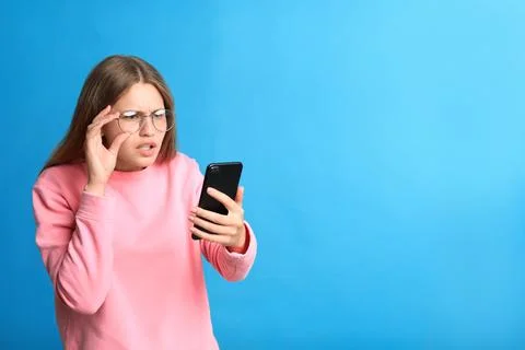 Young woman with vision problems using smartphone on blue background, space f Stock Photos