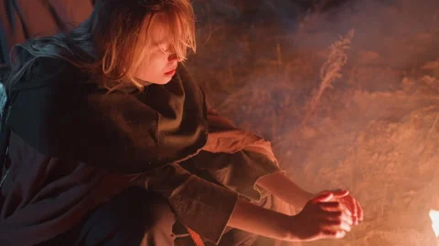 Young woman warming hands over campfire at night in remote outdoor setting with Video stock 343684643