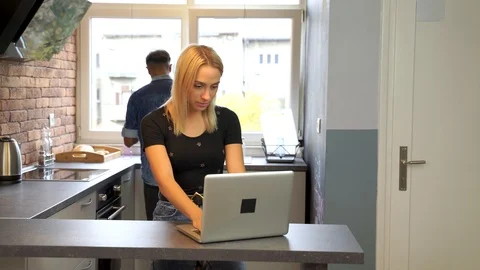 Young woman working at the notebook while boyfriend washes the dishes. Stock Footage 119299986