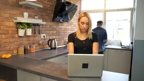 Young woman working at the notebook while boyfriend washes the dishes. Stock Footage 119299996