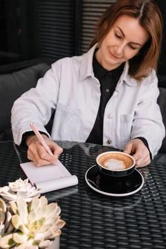 Young woman writes in notebook while enjoying coffee at a cozy cafe in the Stock Photos