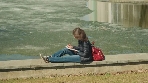 Young Woman Writing in Notebook by Algae Covered Pond with Red Pen and Backpack Stock Footage 345045678