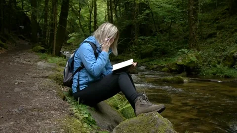 Young woman writing in a notebook, sitting by a forest creek scenic place Stock Footage