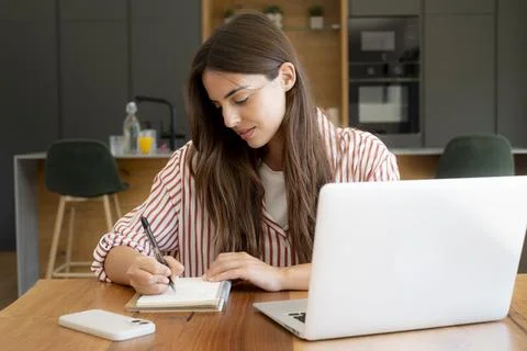 Young woman writing notes while working on laptop in modern kitchen home offi Stock Photos