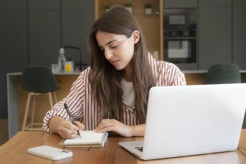 Young woman writing notes while working on a laptop at a kitchen table duri.. Stock-Fotos