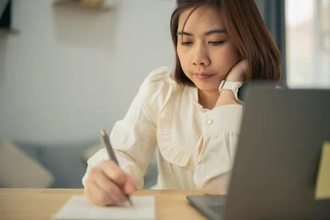 Young woman writing notes while working from home using laptop and pen 库存照片