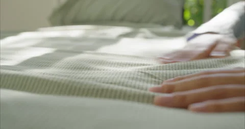 Young woman's hands smooth a striped light green bedcover as morning light glows Stock Footage 335345763