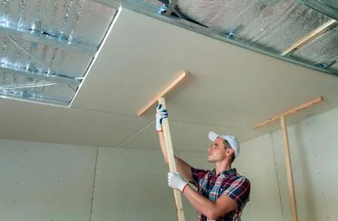 Young worker in protection work gloves fixing wooden holders for drywall susp Stock Photos