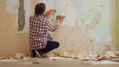 Young worker in protective gloves removing wallpaper from the wall during house Stock Footage 329756647