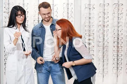 Yung ginger woman enjoying trying on glasses, while optician is tired Stock Photos